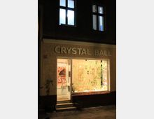Straßenansicht eines Gebäudes mit einem Schaufenster und der Aufschrift „CRYSTAL BALL“ über der Tür. Das Schaufenster ist mit großen, handgeschriebenen Texten und bunten Plakaten dekoriert, und die Fassade ist aus beige verputztem Mauerwerk mit dunkler Fliesenbasis.