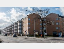 Straßenansicht eines Wohngebäudekomplexes mit abgerundeten Ecken und einer Kombination aus braunen und weißen Fassaden. Entlang der Straße befinden sich parkende Autos, Laternen, ein großer kahler Baum und Geschäfte im Erdgeschoss, darunter ein Reisebüro.