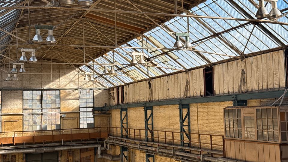 Innenansicht einer ehemaligen Industriehalle mit hohem Glasdach auf Stahlträgern, hängenden Hallenleuchten und großen Sprossenfenstern. Ziegelwände, umlaufende Stahlkonstruktionen und sichtbare Rohrleitungen prägen den Raum.