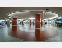 Unterirdischer Durchgangsbereich mit runden Decken- und Bodenmustern, roter und grauer Bodenfliesen, sowie roten Säulen. Im Hintergrund befinden sich Geschäfte, Rolltreppen und Wandwerbungen.