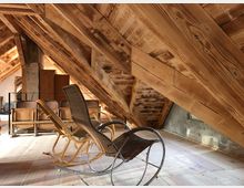 Dachboden mit sichtbarem Holzgebälk und hellen Holzböden. Im Vordergrund stehen ein geflochtener Schaukelstuhl und mehrere Holzstühle, im Hintergrund sind Möbelstücke an einer Wand aus Beton und Holz erkennbar.