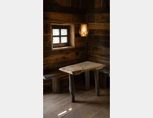 Holzinterieur mit rustikalem Tisch und zwei schlichten Holzbänken in einer Ecke aus Holzpaneelen. Ein kleines Fenster mit mehrfach unterteiltem Glas und eine hängende Lampe mit warmem Licht ergänzen die schlichte, traditionelle Atmosphäre.