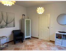 Heller Raum mit Fliesenboden, einer schwarzen Ledersessel-Ecke, einem kleinen, weißen Schrank mit floralem Muster und einer weißen Tür. Eine moderne Deckenlampe hängt an der Decke, ein rundes Wandspiegel und ein Regal mit Büchern und Spielzeug befinden sich an der Wand.