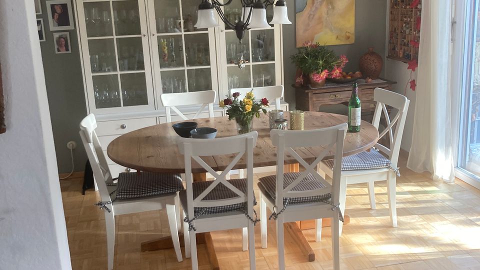 Esszimmer mit einem runden Holztisch, umgeben von weißen Stühlen mit gepolsterten Sitzflächen. Im Hintergrund steht ein weißer Vitrinenschrank vor einer grauen Wand, dekoriert mit Keramikgefäßen und Bildern. Rechts befindet sich eine große Fensterfront mit hellem Tageslicht.