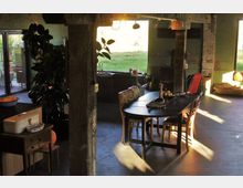 Ein offener Wohnraum mit industriellem Stil, gekennzeichnet durch freiliegende Säulen und Wände mit Sichtmauerwerk. In der Mitte steht ein langer Esstisch mit Stühlen, umgeben von Pflanzen, einem Sofa und großen Fenstern, die Tageslicht hereinlassen.