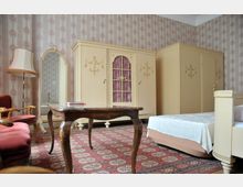 Ein klassisch eingerichtetes Zimmer mit antikem Mobiliar, darunter eine cremefarbene Schrankwand mit goldenen Verzierungen, ein hölzerner Tisch mit geschwungenen Beinen und ein roter Teppich mit Ornament-Muster. Im Raum stehen außerdem ein gepolsterter Sessel, eine große Standspiegel und ein Bett mit weißer Tagesdecke.