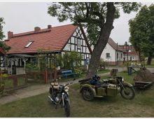 Historisches Fachwerkhaus mit rotem Ziegeldach, umgeben von einem Garten mit Bäumen und Bänken; im Vordergrund stehen zwei Motorräder, eines davon mit Beiwagen.