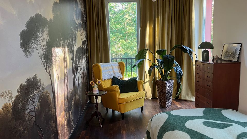 Gemütliches Schlafzimmer mit großer Wald-Fototapete an der linken Wand, dunklem Holzboden und einem gelben Ohrensessel neben einem kleinen Beistelltisch. Vor bodentiefen Fenstern mit goldenen Vorhängen stehen eine hohe Zimmerpflanze und eine Holzkommode; im Vordergrund ist ein Bett mit gemusterter Decke zu sehen.