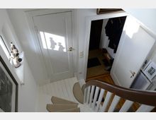 Treppenhaus in einem Wohnhaus mit weiß lackierter Holztreppe und Teppichläufern auf den Stufen. Im Hintergrund eine geschlossene Tür, auf die Sonnenlicht einen Schatten mit Figuren wirft, und eine geöffnete Tür, die in einen Flur mit Holzfußboden führt.