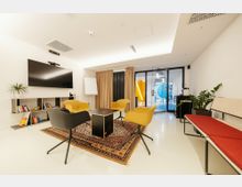 Moderner Besprechungsraum mit weißen Wänden und heller Decke, ausgestattet mit einem großen Bildschirm, einem Flipchart und einem runden Tisch mit gelben und schwarzen Stühlen. Im Hintergrund befinden sich ein Regal mit Büchern, ein Vorhang und eine Glastür, die in einen Flur mit bunten Elementen führt.