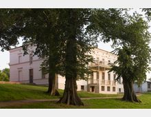Historische Villa mit weißer Fassade und verziertem Balkon, umgeben von großen Bäumen und einer Rasenfläche. Im Hintergrund ist ein weiteres Gebäude zu sehen.