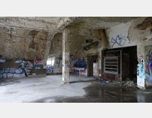 Verlassener Raum mit stark beschädigten Wänden, abblätternder Farbe und Graffiti. Der Raum hat eine Betonstruktur mit sichtbaren Rohren, Fliesen an den unteren Wänden und Wasserlachen auf dem Boden.