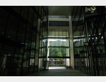 Moderne Glaspassage mit hohen, spiegelnden Fensterfronten auf beiden Seiten, die in einen offenen Innenhof mit Blick auf ein Gebäude und Bäume führt. Fahrräder stehen am Rand der Passage, und ein Bistro-Schild ist sichtbar.