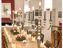 Ein festlich gedeckter langer Holztisch in einem Innenraum mit weißen Servietten, grünen Adventskränzen mit roten Kerzen und silbernen Kerzenleuchtern mit brennenden, dunkelgrünen Kerzen. Im Hintergrund sind rote und weiße Wände sowie gerahmte Bilder zu sehen.