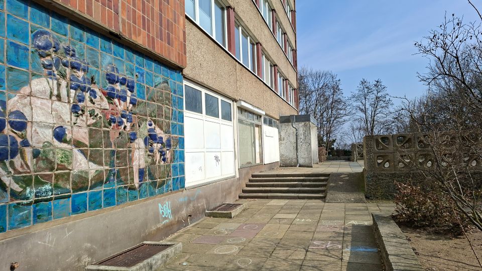 Außenansicht eines mehrstöckigen Wohn- oder Bürogebäudes mit verputzter Fassade und einem großen Wandmosaik aus blauen Fliesen. Davor liegt ein gepflasterter Gehweg mit Stufen und niedriger Betonbrüstung, umgeben von kahlen Bäumen und Sträuchern.