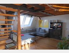 Wohnzimmer in einer Dachgeschosswohnung mit sichtbaren Holzträgern und Holztreppe. Die Einrichtung umfasst ein großes graues Ecksofa, einen kleinen Glastisch, einen antiken Holzschrank und helle Dachfenster, durch die Tageslicht einfällt.