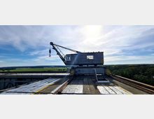 Ein alter, hölzerner Kran mit einem Ausleger und Haken steht auf Schienen auf einer erhöhten Metallplattform. Im Hintergrund erstreckt sich eine Landschaft mit Wiesen, Wäldern und einem klaren Himmel bei Sonnenlicht.