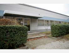 Flacher Gebäudeeingang mit verglaster Tür und Vordach, umgeben von Hecken und kahlen Sträuchern. Die Fassade ist in hellen Farben gehalten, mit einem langen Fensterstreifen, dessen Jalousien teilweise geschlossen sind.