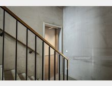 Treppenhaus mit einer Wand aus Sichtbeton, einem Geländer mit schwarzen Streben und einem hölzernen Handlauf. Im Hintergrund ist eine helle Türöffnung zu sehen, die in einen angrenzenden Raum führt.