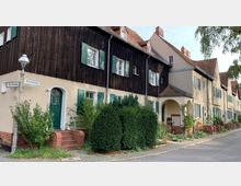 Reihenhäuser aus einer historischen Arbeitersiedlung mit verputzten Fassaden und Holzverkleidungen, grün gestrichenen Fensterläden und roten Ziegeldächern; vor den Häusern befinden sich Vorgärten mit Hecken und Blumen. Straßenschilder weisen die Wege „Buchenweg“ und „Eichenweg“.