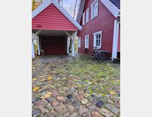 Rotes Holzhaus mit weißen Fensterrahmen und einem überdachten Carport auf der linken Seite, umgeben von einem gepflasterten Hof mit moosbewachsenen Steinen und herabgefallenen Herbstblättern. Ein Fahrrad lehnt an der Seitenwand des Hauses.