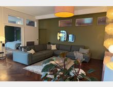 Wohnzimmer mit grüner Wand, großem grauen Ecksofa und runden Spiegeln an der Wand. Im Vordergrund ein Teppich mit Couchtisch und Pflanzen, links ein Durchgang mit Blick in ein Schlafzimmer, rechts moderne Beleuchtung und Holzfußboden im Fischgrätmuster.