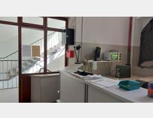 Ein Büro- oder Arbeitsraum mit einem Schreibtisch, auf dem Unterlagen, Büroutensilien, ein Monitor und eine Schreibtischlampe stehen. Im Hintergrund ist eine Glastür, die zu einem Treppenhaus mit Betonstufen und Metallgeländern führt.