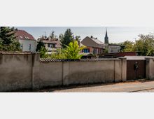 Straßenszene mit einer niedrigen, verputzten Mauer im Vordergrund, dahinter Wohnhäuser mit roten und grauen Dächern sowie Bäumen. Im Hintergrund ist der Turm einer Kirche zu sehen.