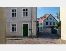 Straßenszene in einer Altstadt mit Kopfsteinpflaster, einem einfachen zweistöckigen Haus mit grünem Eingang und vier Fenstern links, und einem größeren Fachwerkhaus mit rotem Ziegeldach im Hintergrund. Rechts ist eine Ziegelsteinmauer mit einem geschlossenen Tor zu sehen.