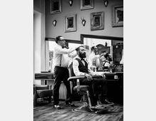 Schwarz-weiß-Foto eines Frisörsalons mit Retro-Einrichtung. Ein Mann mit Bart sitzt auf einem Frisierstuhl, während ein Friseur im Hemd und Hosenträgern ihm die Haare schneidet; im Hintergrund ein großer Spiegel und eingerahmte Bilder an der Wand.