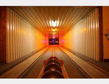 Ein schmaler, länglicher Kegelbahnraum mit maschinellen Kegelaufstellern am Ende der Bahn. Die Wände und Decke sind aus hellem, geripptem Material, und warme gelbliche Beleuchtung erhellt den Raum. Im Vordergrund liegen mehrere schwarze Kegelkugeln.