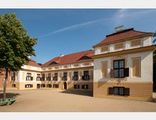 Barockgebäude mit cremefarbener Fassade und gelben Akzenten, dunklen Fensterläden und rotem Ziegeldach. Im Vordergrund ein sandiger Platz und ein einzelner Baum.