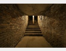 Treppenaufgang in einem engen unterirdischen Gang mit Wänden aus rohen Backsteinen und schwacher Beleuchtung. Die Treppe führt zu einer Öffnung mit Tageslichteinfall.