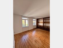 Helles Zimmer mit Holzdielenboden, zwei großen Fenstern mit Blick nach draußen und einer Wand mit eingebautem Regalsystem aus Holz. Die Wände sind weiß gestrichen, und ein Heizkörper befindet sich unter einem der Fenster.