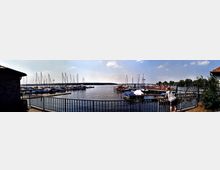 Panorama eines Hafens mit zahlreichen Segelbooten und kleinen Motorbooten, die an Stegen vertäut sind. Im Vordergrund ein Metallgeländer und eine Person, die auf das Wasser blickt, im Hintergrund Baumreihen und ein heller Himmel.