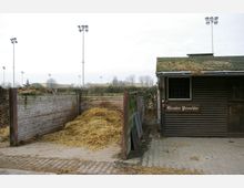 Ein Außenbereich mit einem Holzverschlag, in dem eine große Menge Stroh gelagert ist, sowie einem kleinen Schuppen mit der Aufschrift „Günter Verschke“. Im Hintergrund sind Flutlichtmasten und Bäume sichtbar.