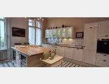 Helle, klassische Küche mit weißen Einbauschränken, Holz-Arbeitsflächen und einer langen Kücheninsel; an der Rückwand ein blau-weiß gefliestes Muster mit dekorativem Motiv. Große Fenster bringen viel Tageslicht, und der Boden ist in einem schwarz-weißen geometrischen Fliesenmuster gehalten.