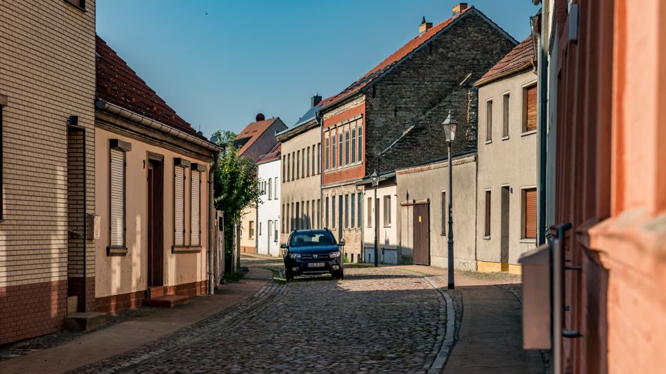 Schmale Kopfsteinpflasterstraße in einem Wohngebiet mit dicht stehenden, niedrigen Häusern mit Putz- und Ziegelfassaden sowie roten Satteldächern. Entlang der Straße stehen eine Laterne und ein geparktes Auto, im Hintergrund setzt sich die Häuserzeile fort.