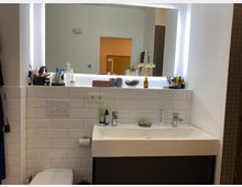 Badezimmer mit weiß gefliester Wand und einem großen beleuchteten Wandspiegel. Ein weißes Waschbecken mit verschiedenen Pflegeprodukten steht unter dem Spiegel, daneben eine Toilette mit Aufputz-Spültasten. Im Hintergrund ist eine geöffnete Tür und ein braun gefliester Duschbereich erkennbar.