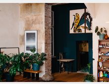 Innenraum einer modern eingerichteten Wohnung mit sichtbaren Texturen wie unverputzter Ziegelwand und blasser Wandfarbe. Im Vordergrund stehen Zimmerpflanzen in Töpfen, daneben ein Holzhocker, während an den Wänden Kunstwerke und ein Bücherregal mit Farben und Deko-Objekten hängen. Rechts ist ein kleiner Durchgang zu einer weiteren Nische erkennbar.