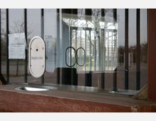 Ein geschlossenes Kassenhäuschen mit Glasscheiben, durch die ein leerer Außenbereich mit Bäumen und Gebäuden im Hintergrund sichtbar ist. An der Scheibe hängt ein Zettel mit Eintrittspreisen, daneben ein Schild mit der Aufschrift "Geschlossen".