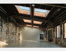 Ein großer, leerer Industrie-Raum mit Betonboden, gealterten, teilweise verputzten Ziegelwänden und einer Decke mit mehreren Oberlichtern aus Glas. Die Fenster sind hoch und in Metallrahmen gefasst, die Beleuchtung erfolgt über an der Decke montierte Schienenscheinwerfer.