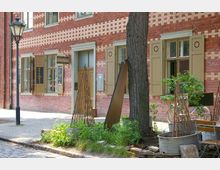 Backsteinfassade eines Gebäudes mit hellbraunen Fensterläden und der Beschilderung „WeinWerk“. Im Vordergrund stehen hölzerne Pflanzenstützen in Metallkübeln, ein Baum und ein gepflasterter Gehweg mit einer Straßenlaterne.