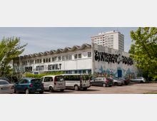 Ein flaches Gebäude mit wellenförmigem Dach und Graffiti-bemalten Wänden, darunter die Schriftzüge "Power statt Gewalt" und "Einheit statt Gewalt". Im Vordergrund befindet sich ein Parkplatz mit mehreren geparkten Autos, während im Hintergrund ein Hochhaus und Bäume zu sehen sind.