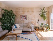 Wohnzimmer mit heller, minimalistischer Einrichtung: Ein beiger Stoffsofa steht vor einer unbehandelten, beige-braunen Wand mit Kunstwerk. Pflanzen wie Monstera und eine große Grünpflanze schmücken den Raum. Auf einem Holzboden liegt ein Teppich mit buntem Muster, neben einem hellen Couchtisch.