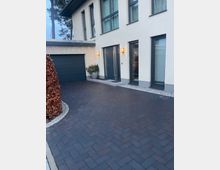 Moderne Hausfassade mit heller Putzoberfläche und großen Fenstern, daneben eine integrierte Garage mit dunkelgrauem Tor. Der Eingangsbereich ist mit einer Tür in anthrazitfarbenem Rahmen und Außenbeleuchtung gestaltet. Davor liegt eine gepflasterte Einfahrt.