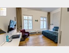 Ein modernes Hotelzimmer mit einem großen Doppelbett, blauem gemusterten Bettbezug, Holzboden und weißen Wänden. Zwei Fenster mit Vorhängen lassen Tageslicht herein; links steht ein Schreibtisch mit Stuhl, Wasserflasche und Wandfernseher, daneben ein roter Sessel.