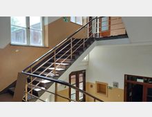 Treppenhaus in einem Gebäude mit hell gestrichenen Wänden und braun lackierten Metallgeländern. Große Fenster lassen Tageslicht herein, und ein Fluchtweg-Schild zeigt Richtung Ausgang, der unten zu einer Holztür mit Glaseinsätzen führt.