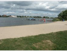Ein sandiger Strandbereich an einem See mit einer Badezone, die durch eine Kette mit orangefarbenen Bojen abgegrenzt ist. Im Wasser steht ein Steg mit einer roten Rutsche, im Hintergrund sind Gebäude und Bäume am anderen Ufer zu sehen.