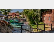 Flusslandschaft mit Anlegestelle für grün abgedeckte Boote, umgeben von Gebäuden aus rotem Backstein und Bäumen. Im Hintergrund ist ein historisches Haus mit Satteldach zu sehen, umgeben von einer Uferpromenade mit Sitzbereichen.